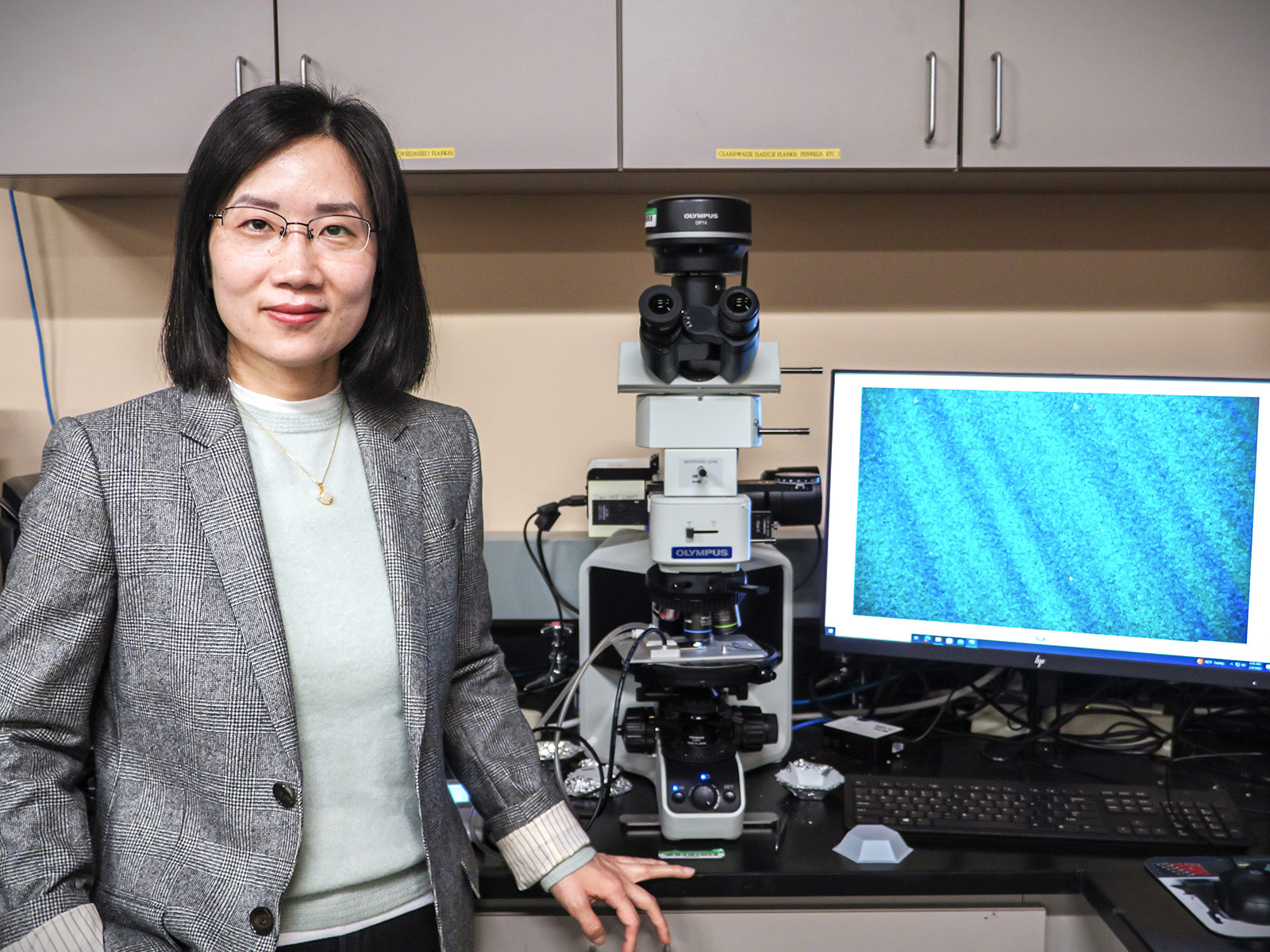 A woman in a gray blazer and glasses poses, smiling. She stands next to a microscope and computer monitor.