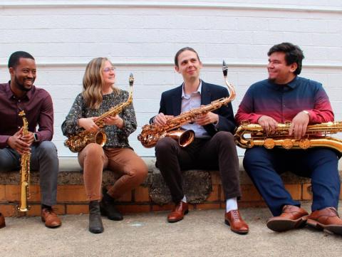 Lotus Saxophone Quartet members Rico Allen, Mikayla Peterson ('22 M.M.) , Trek Boyland and Ben Facundo sitting on a curb holding their instruments.