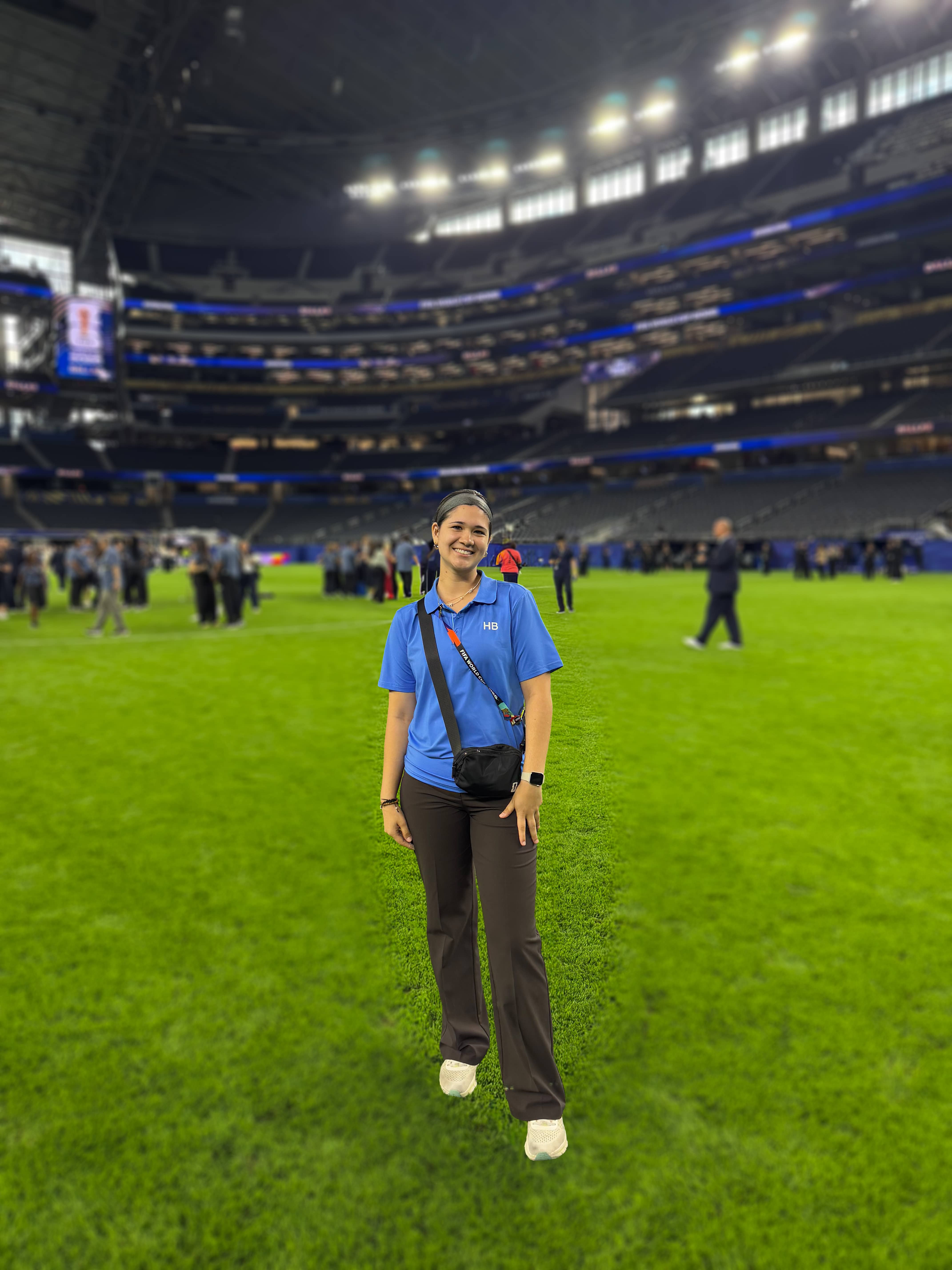 A young woman in a blue polo shirt stands on the turf in a stadium that's blurred