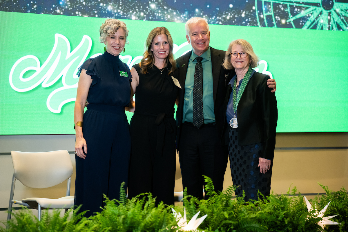Three women and one man stand side by side posing for a photo. The man wears a green shirt. The woman on the far right, Dornith Doherty, wears an medal for her award