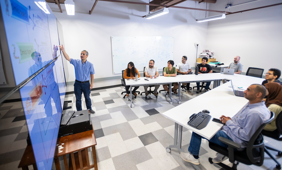 A man in a blue button down and navy pants stands at a screen displaying information. Students sitting along tables watch him.