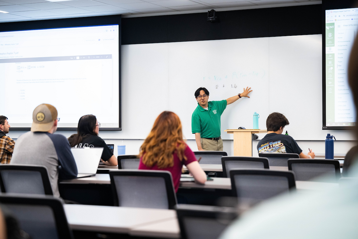 A man in a green shirt stands before a class and references text on the white board behind him.