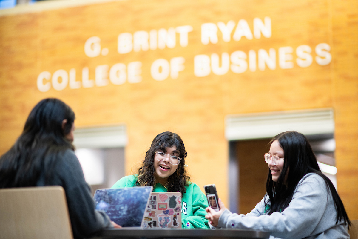 A group of three young women sit at a table working on laptops and a phone. Two are smiling while a third has her back to the camera. Letting on the wall behind them reads "G. Brint Ryan College of Business."