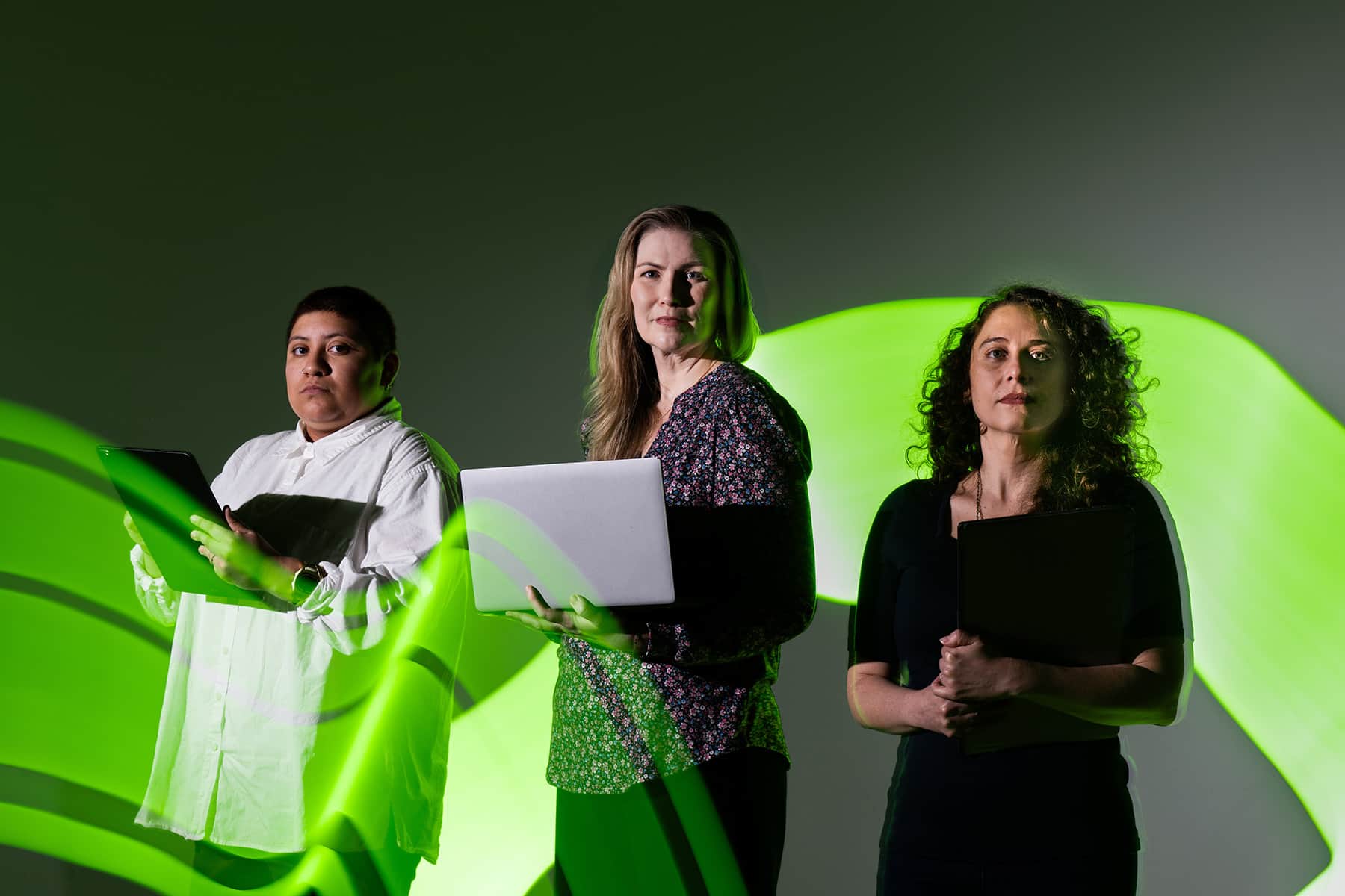Three people stand holding laptops and electronic devices while a green ribbon of light weaves in front of and behind them.