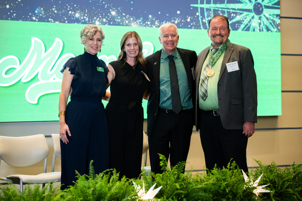 Two women and 2 men stand side by side posing for a photo. The women wear all black while the men wear green shirts with black pants and blazer. The man on the far right, Paul Hensel, wears an award medal