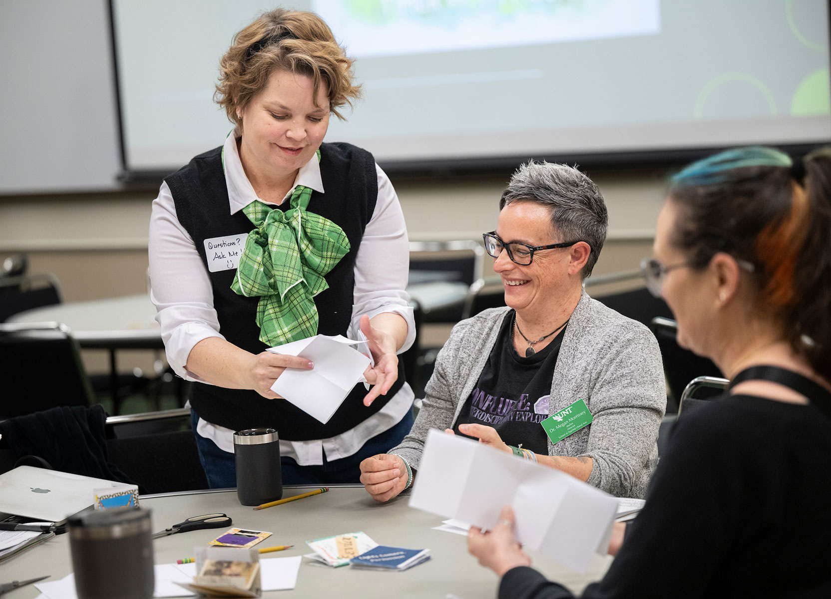 A woman in a white shirt, black vest and green tie speaks to a woman in a gray sweater. They are smiling.