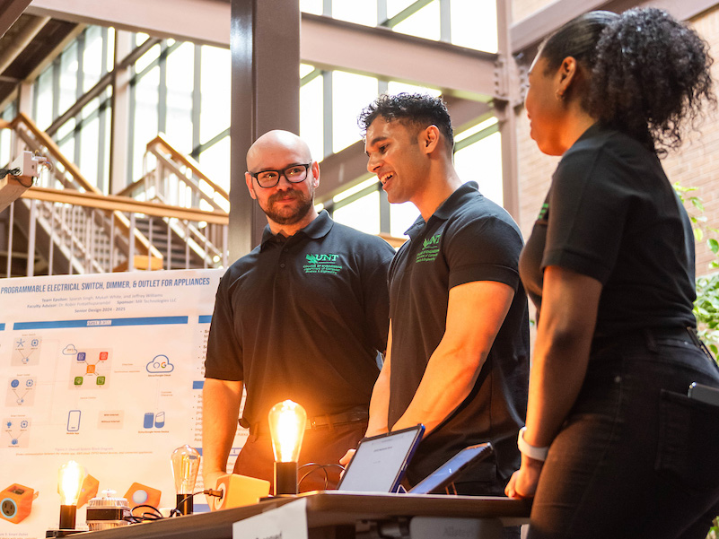 Three student stand at a table facing left, on the table are 2 lit up lightbulbs 