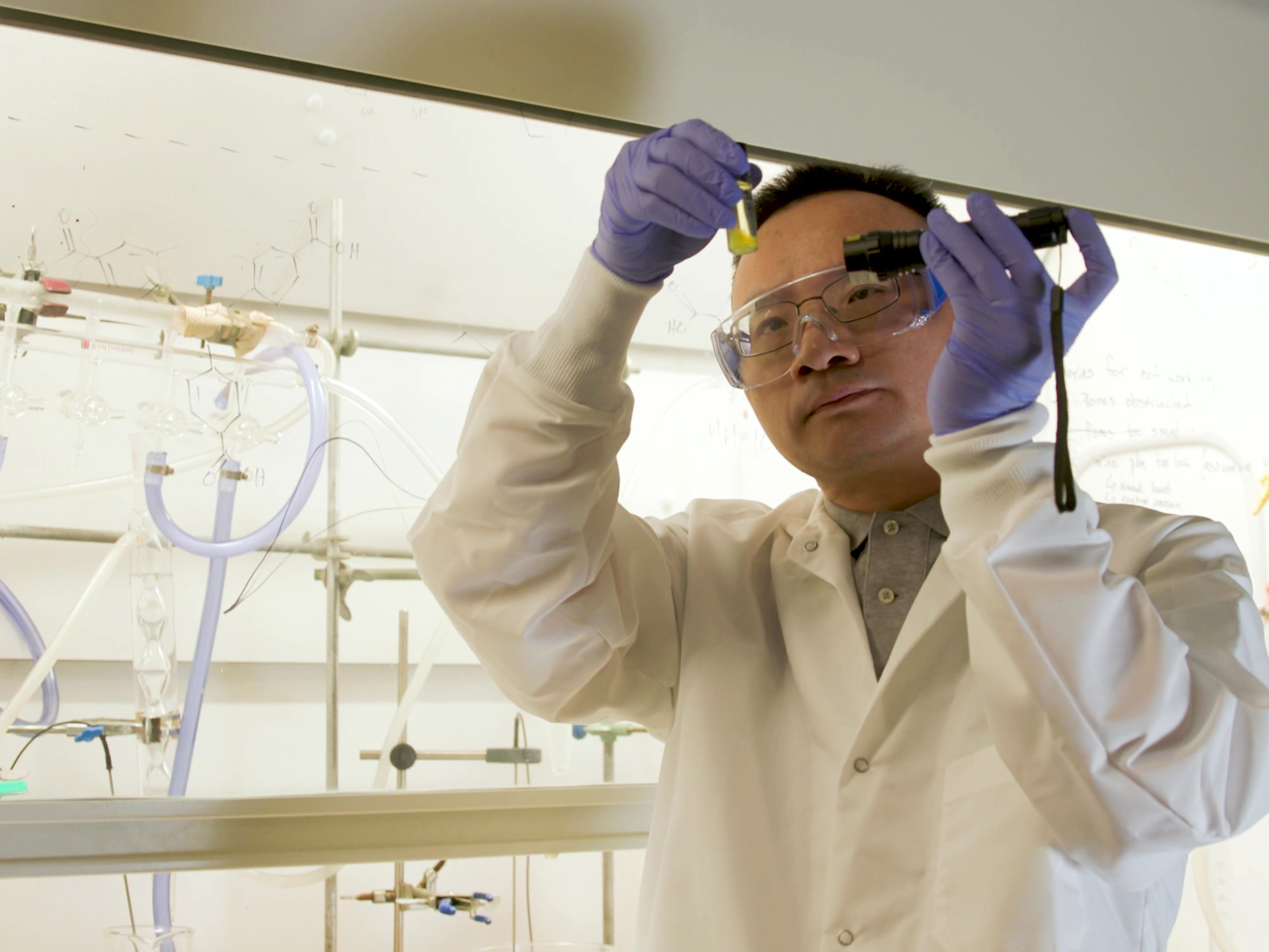 A man in a lab coat, safety goggles, and gloves looks at a vial with a liquid sample in it. He holds a light to the vial. He stands in a lab.