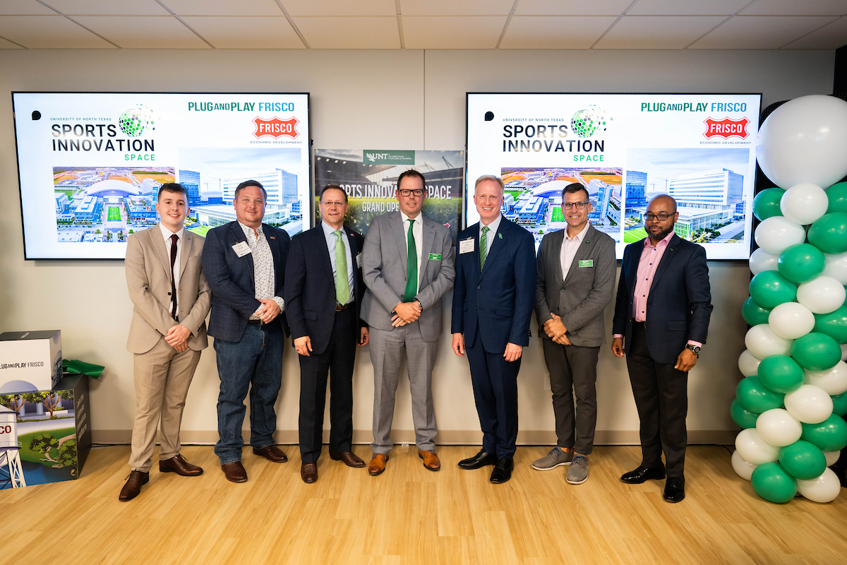 Seven men stand together in front of screens that say Sports Innovation with the Frisco logo in the corner