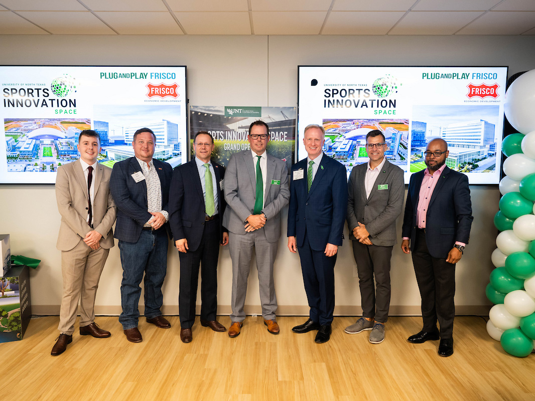 Seven men in suits stand posed for a photo. Behind them are two large screens that say Sports Innovation space. Green and white balloons are to the right of them