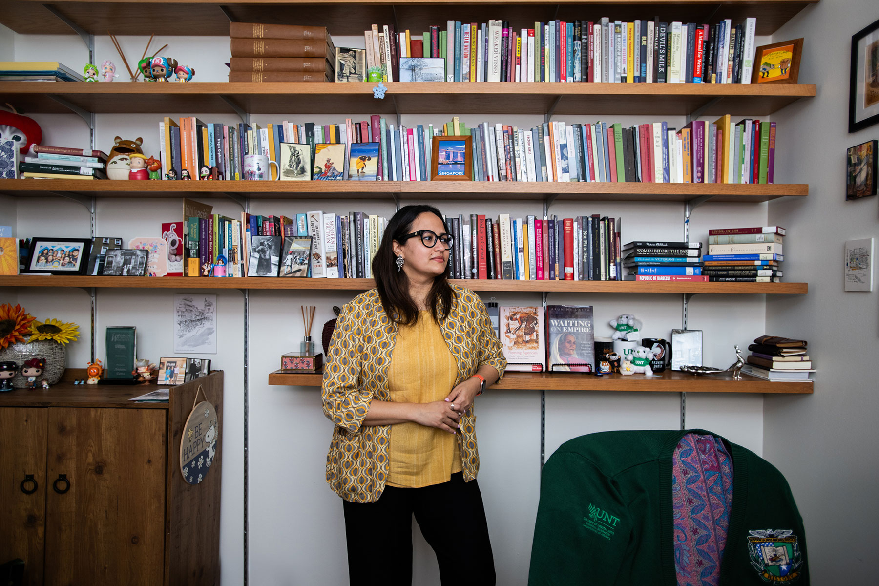 A woman in a yellow blouse and overshirt poses in front of four shelves lined with books and action figures