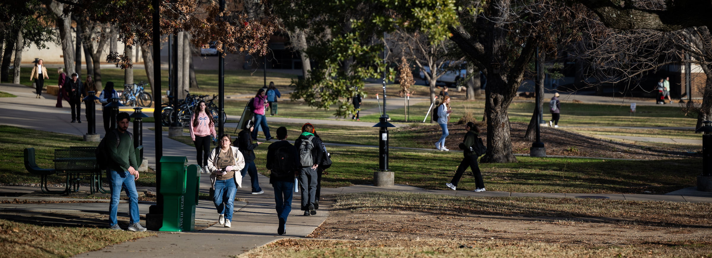 Students walk on campus in a central area with many sidewalks and full grown trees.