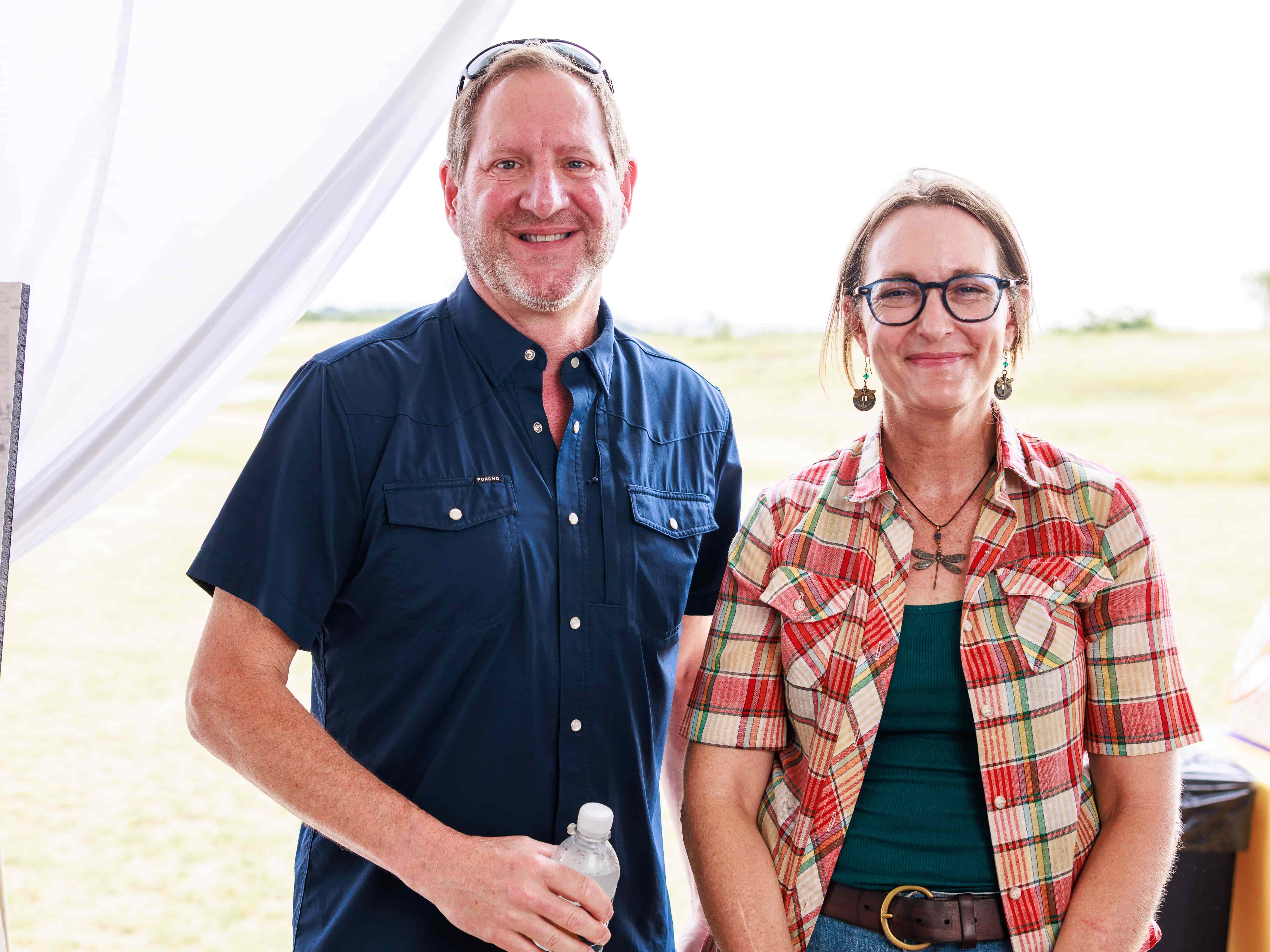 A man in a dark blue button down stands next to a woman with glasses  in a plaid over shirt and blue undershirt.