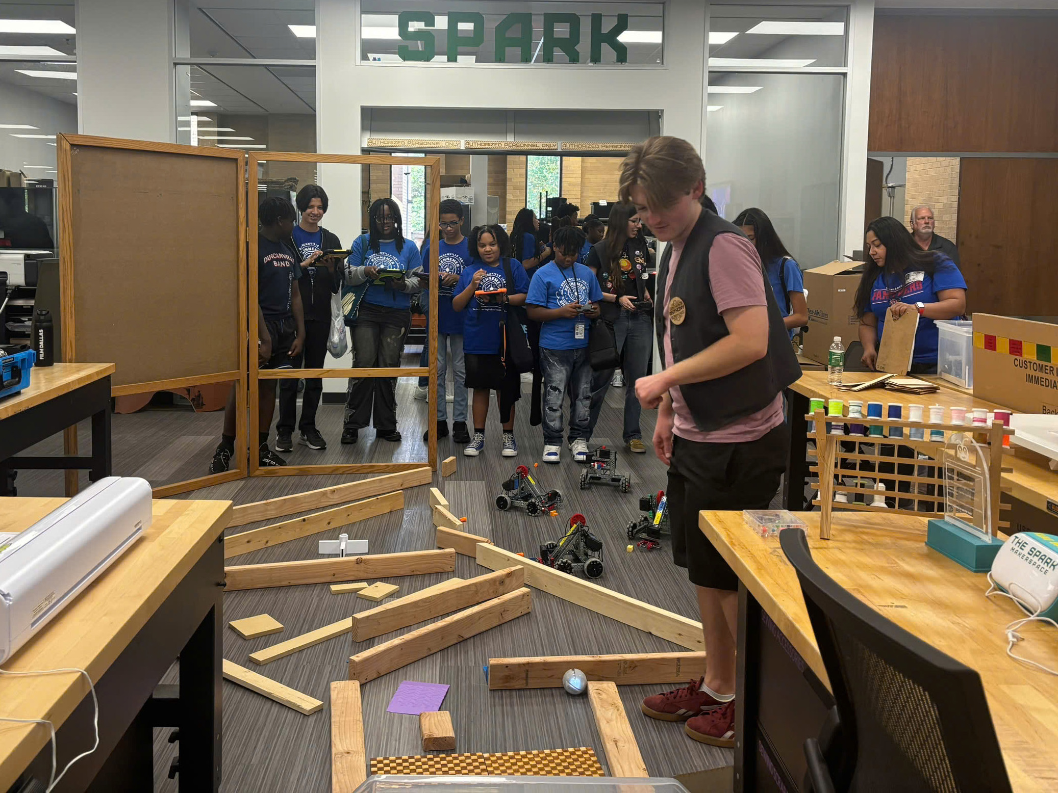 A group of student sin blue stand in the background. The hold control panels for machines going through a makeshift obstacle course on the floor in front of them. A library worker stands in the foreground watching the machines.