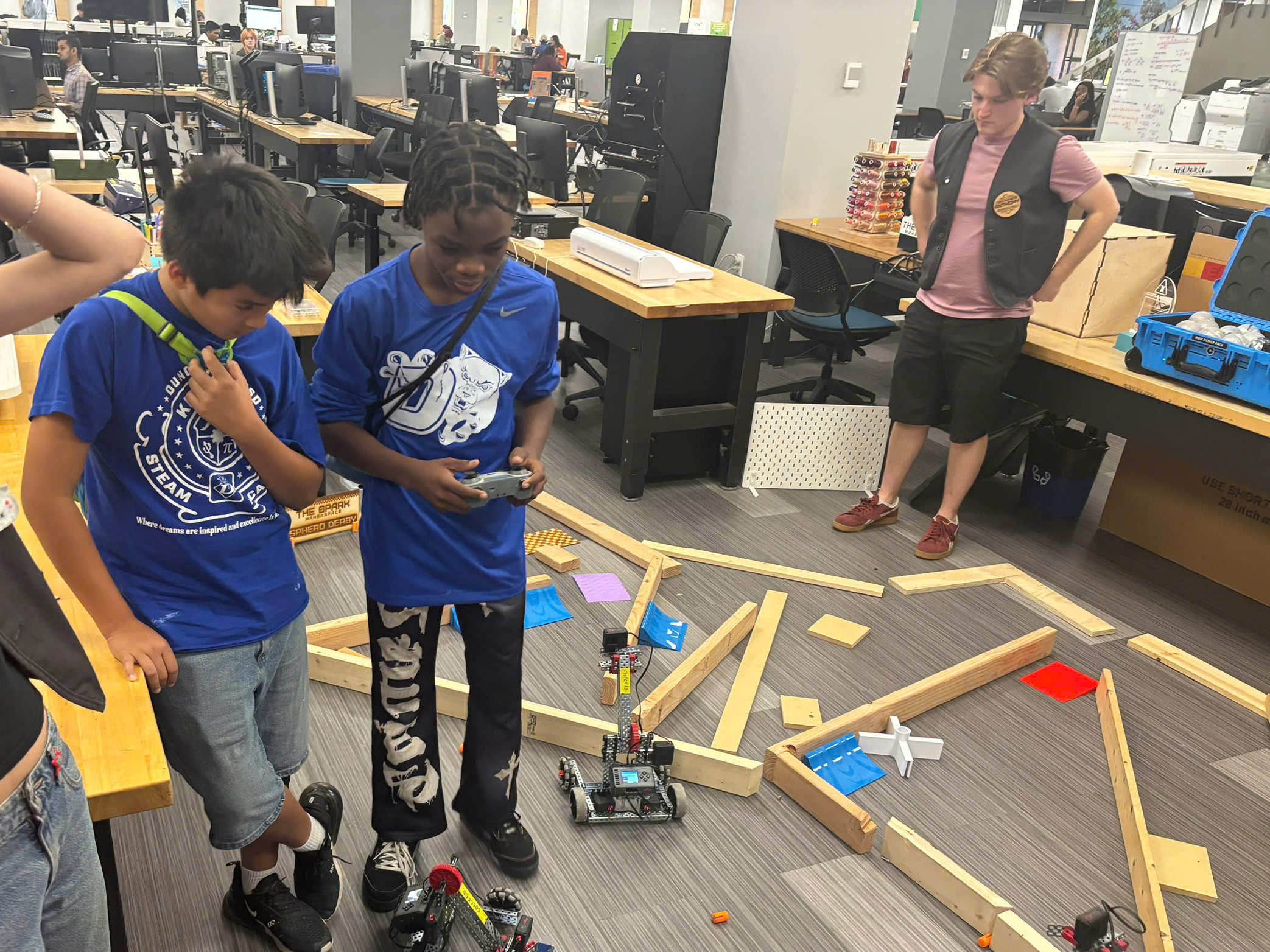 Two middle school boys in blue shirts look down at a machine one if controlling. To the right is an obstacle course for the machine. A library worker stands in the background.
