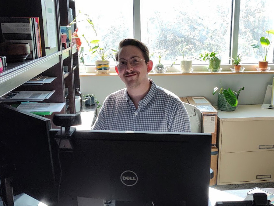 A man with glasses sits at a desk behind a computer. There is a window behind him with multiple plants on the windowsill