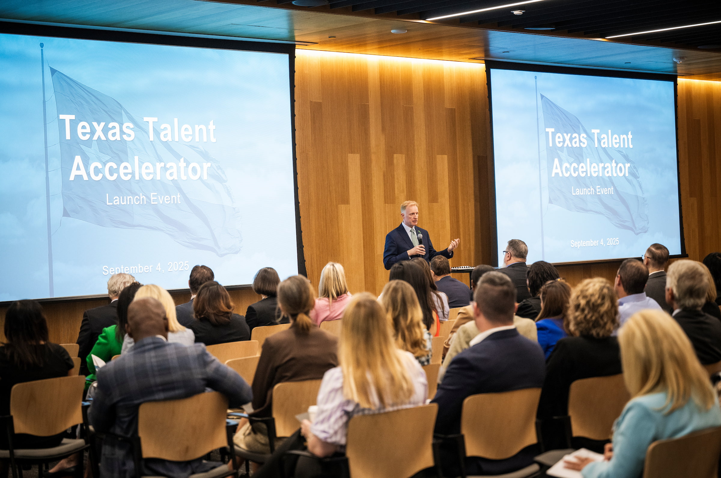 President Keller speaks in the center of a room to a group of people sitting down facing him. He is flanked by two screens that say "Texas Talent Accelerator"