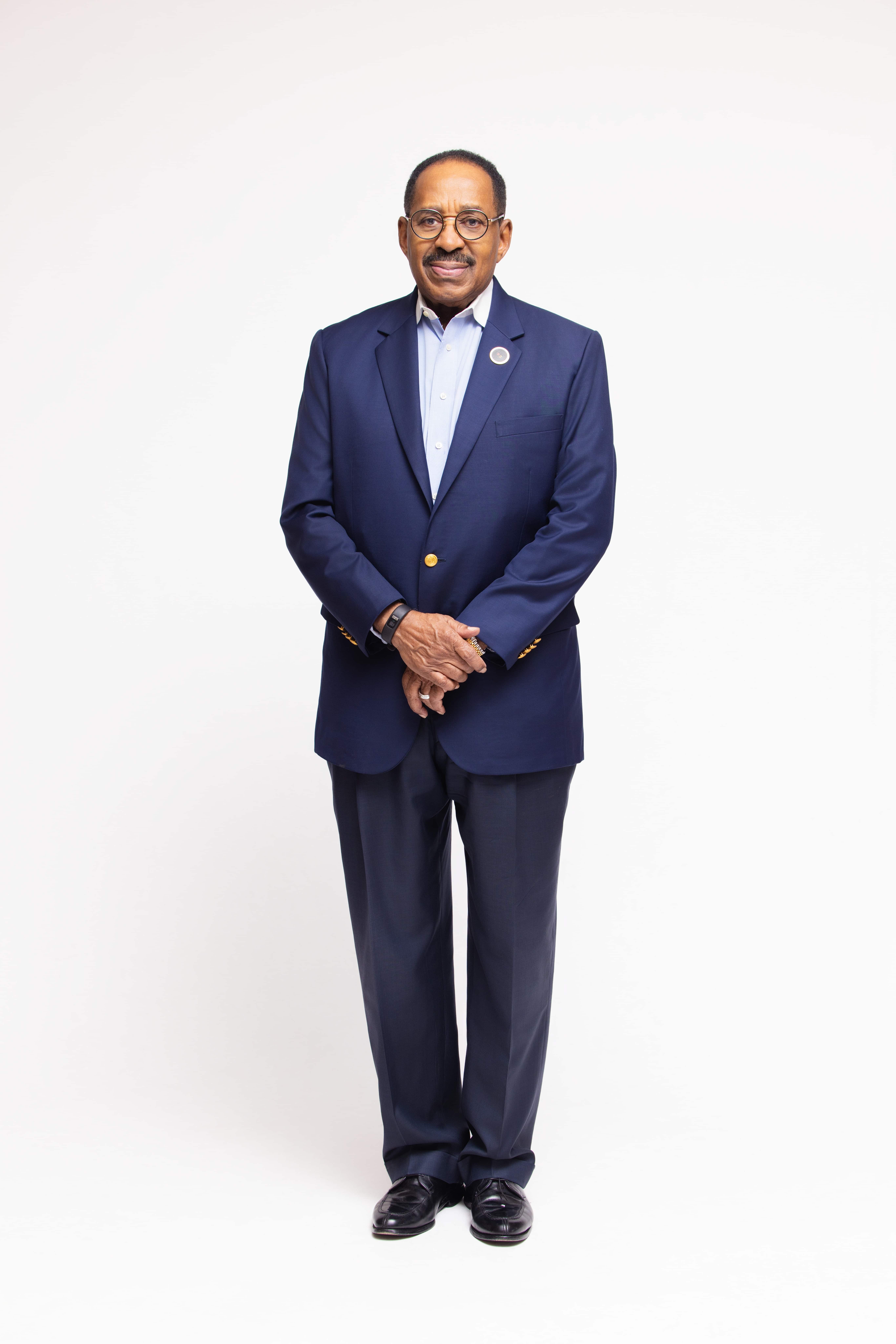 Levi H. Davis stands in front of a white background in a navy blue suit with his hands clasped together, he wears glasses