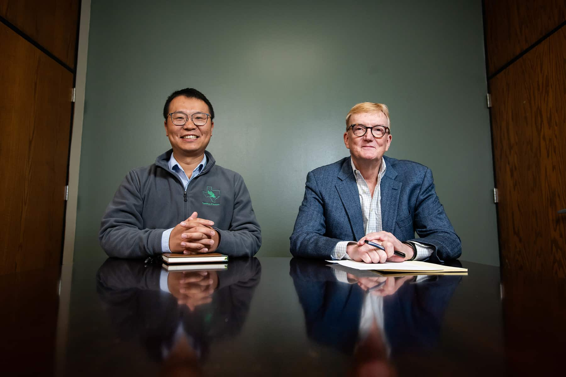 Dr. Yang Zhou and Dr. Michael Carroll sit side by side at a table in front of a green background