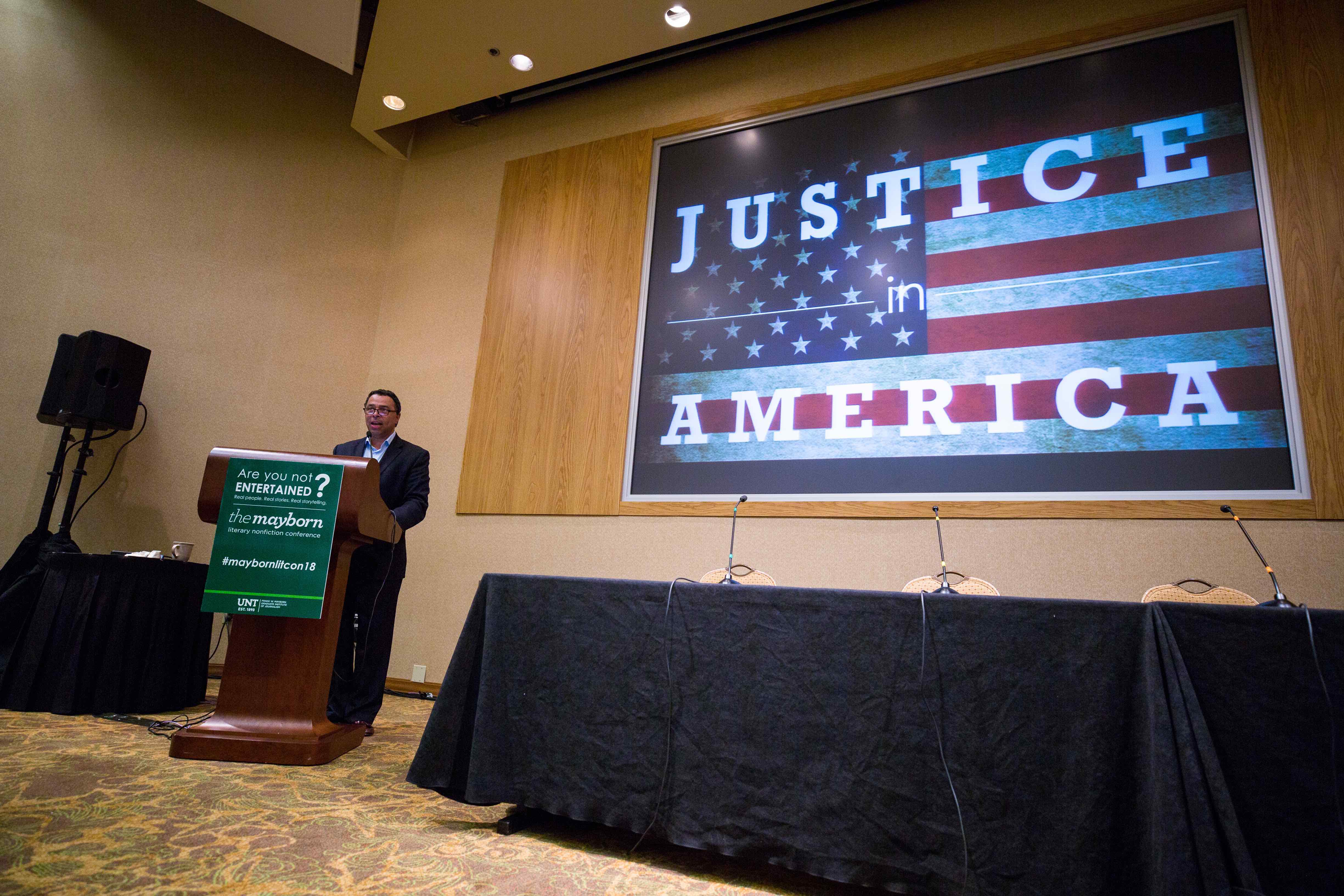 2018 Mayborn Conferen organizer Neil Foote stands next to a power point/banner presenting the (now) current theme: Justice in America