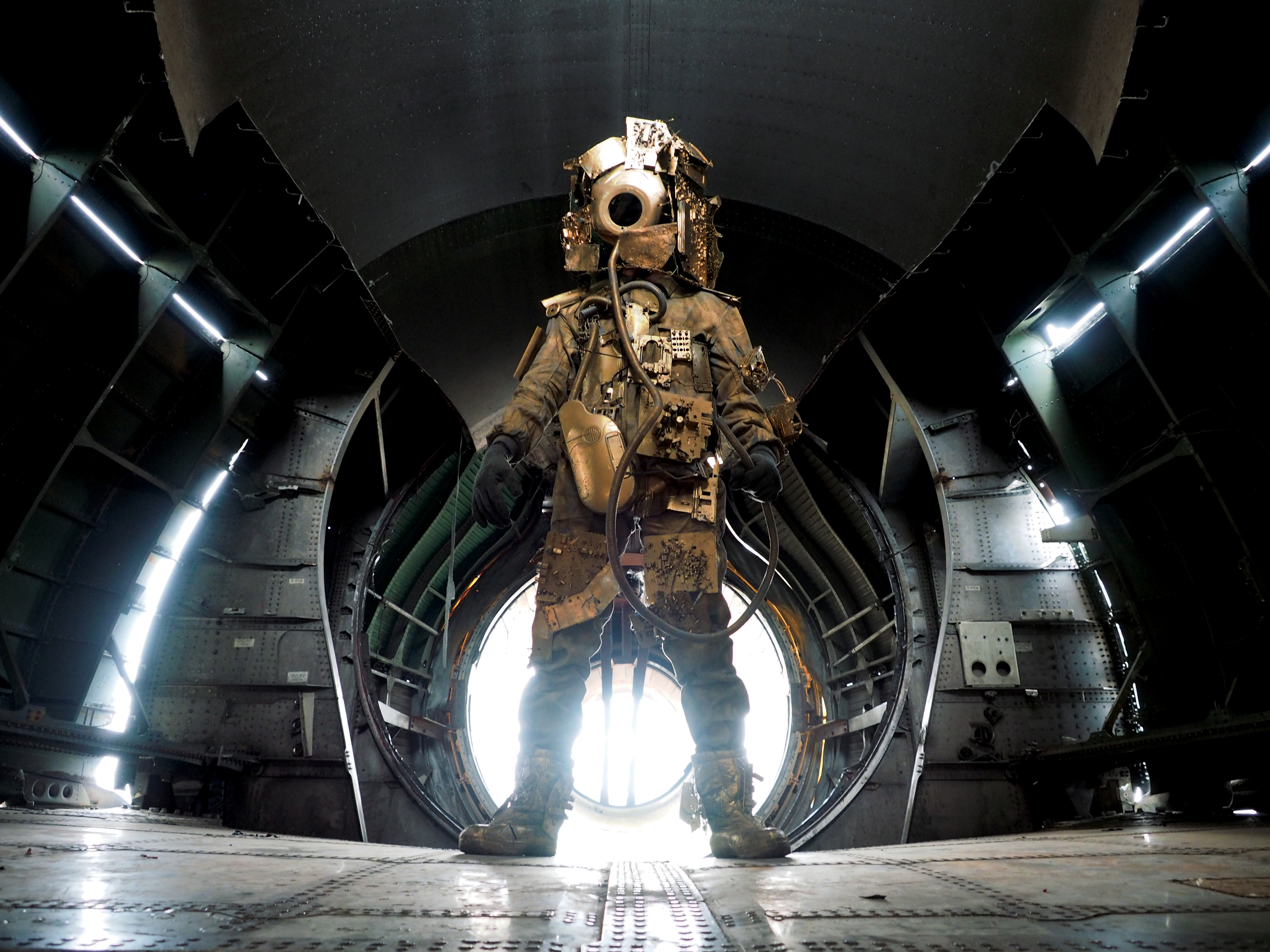 Photo of a humanoid figure dressed in a gold metallic makeshift space suit and helmet standing in the body of a gutted airplane showing the metal bracing and studs with a circle of light behind them.