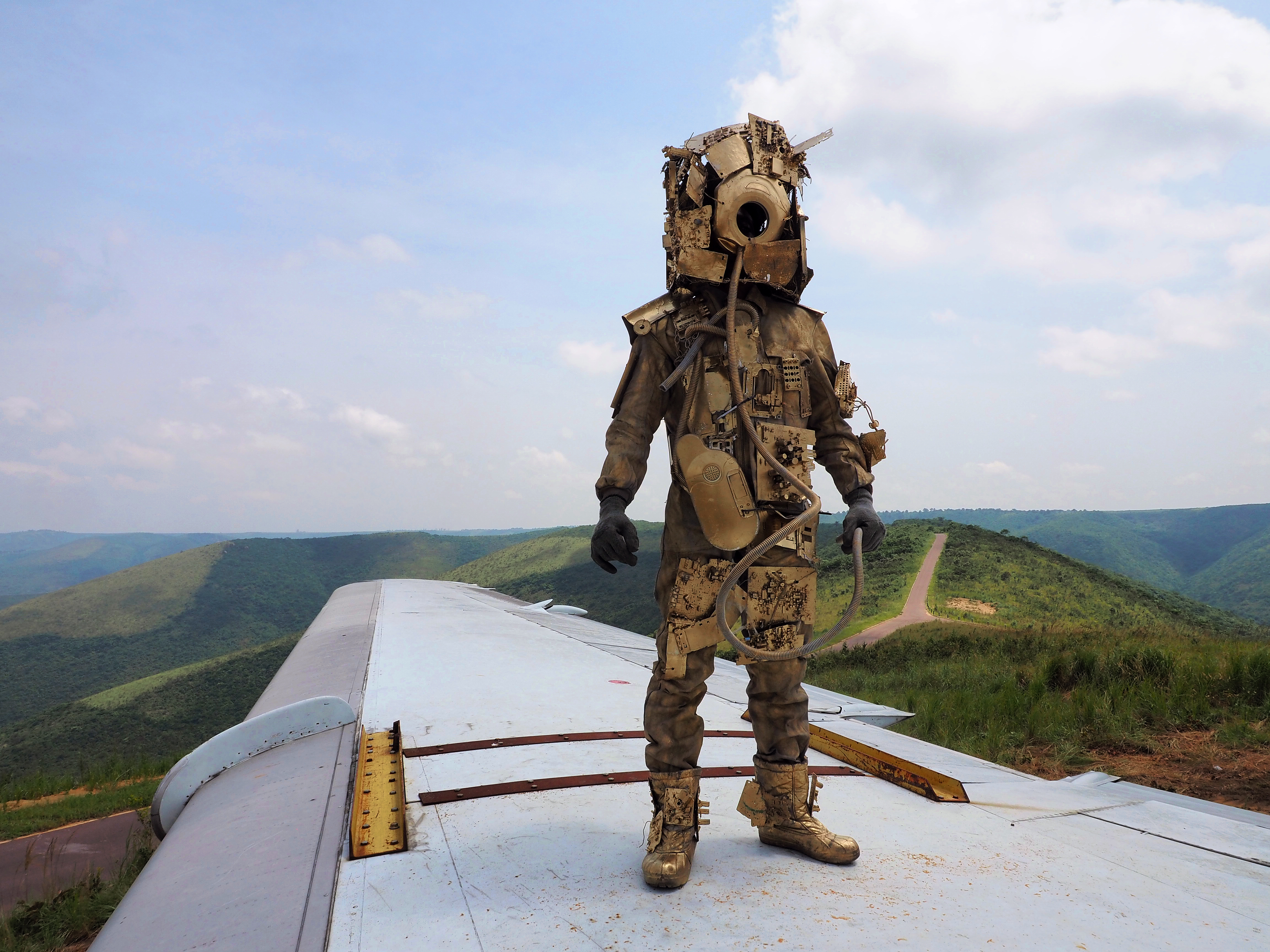 A humanoid figure in a gold metallic makeshift space suit and helmet stands on an airplane's wing with a landscape beyond. Green rolling hills and paths can be seen with a slightly blue cloudy sky in the image's background.
