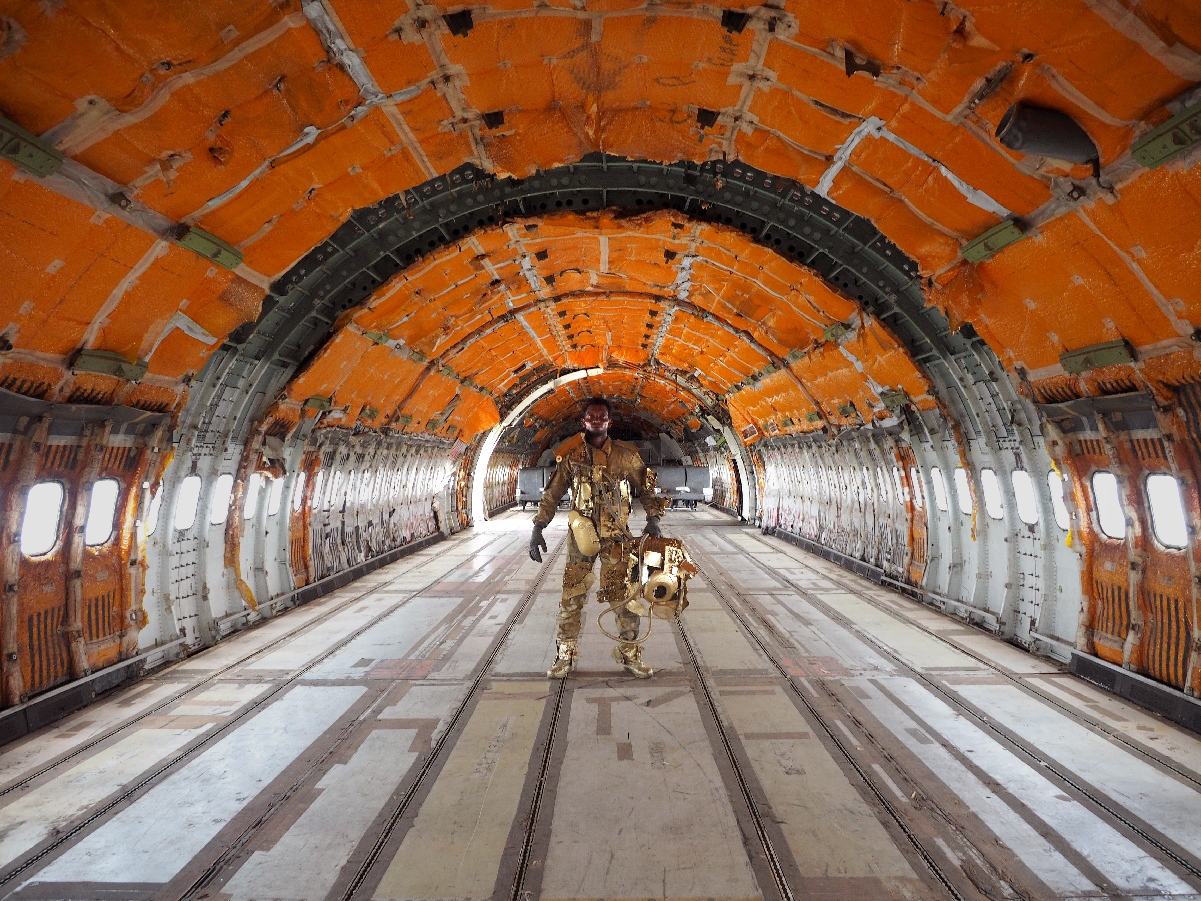 In a gold metallic makeshift space suit holding an astronaut helmet, a Black man is standing in the middle of an empty cargo plane of receding orange arches.
